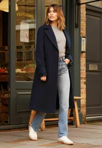 Morski dugi kaput sa širokim reverima, sivi pulover i svijetloplave trapezice. Bijele tenisice s gumiranim đonom. Stoji blizu ulaza u trgovinu.