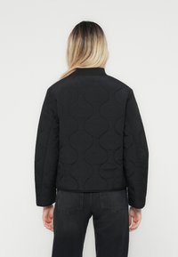 Veste matelassée noire avec un motif de vagues texturé, col côtelé et manches longues, associée à un jean foncé. Design simple et minimaliste.