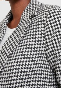 Blazer a pied-de-poule in bianco e nero, con tessuto strutturato, colletto a lancia e vestibilità sartoriale, che mette in risalto il design del motivo intricato.