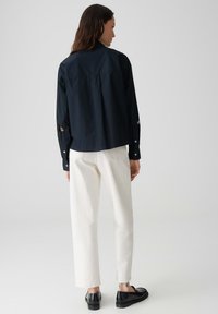 Navy shirt with a boxy design, long sleeves, and button cuffs; paired with white straight-leg trousers and black slip-on shoes.