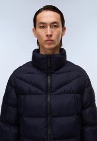 Chaqueta de plumas azul marino con cuello alto, cierre de cremallera y diseño acolchado. Presenta una textura suave y detalles de cremallera negra en contraste.