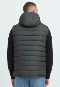 Halloev gray puffer vest, millel on tikitud tekstuur, kõrge krae ja kapuuts. Kandmine musta sviitri peal, näidates lõdva sobivuse ja kihilise disaini.