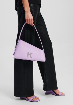 Lila handtas met een driehoekige vorm en een zilveren logo-detail. Gemaakt van soepel leer en heeft een enkele handgreep. Draagt bij zwarte wijde broek.