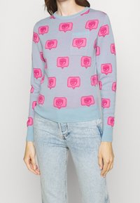 Ljusblå tröja med rosa grafiskt mönster. Har rund halsringning, långa ärmar och en bröstficka. Stickad tyg med mjuk textur.