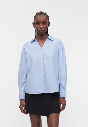 Žena s vrkočmi má na sebe voľnú svetlomodrú košeľu na gombíky a čiernu asymetrickú minisukňu na jednofarebnom pozadí.