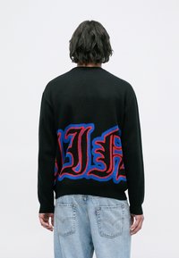 Maglione nero lavorato a maglia con audace scritta grafica rossa e blu sul retro, con polsini e orlo a costine. Indossato con jeans in denim chiaro.