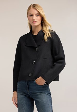 Donna bionda che indossa un cappotto nero abbottonato e jeans blu, in piedi con una mano in tasca davanti a uno sfondo chiaro e semplice.