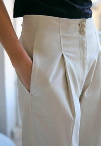 Pantalons taille haute beige clair avec deux plis à l'avant, poches latérales et ceinture à boutons comportant deux boutons, fabriqués en tissu lisse.