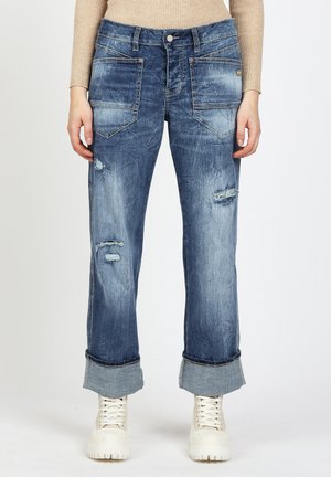Person wearing cuffed, distressed blue jeans with large front pockets and off-white chunky sneakers, standing against a plain background.