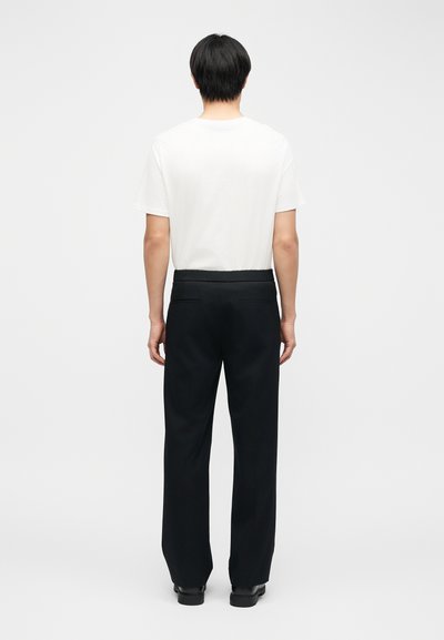 Άνδρας που φοράει απλό λευκό κοντομάνικο μπλουζάκι, μαύρο παντελόνι ίσιο, και μαύρα παπούτσια, στέκεται με την πλάτη γυρισμένη σε λευκό φόντο.