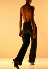 Žena odchádzajúca, oblečená v tmavých vysokých džínoch, čiernom halterneck top neotvorenom na chrbte a čiernych topánkach na vysokom opätku, na béžovom pozadí.