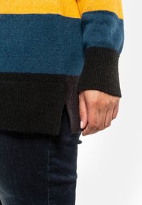 Pull en tricot avec de larges rayures jaunes, bleu sarcelle et noires. Il présente des poignets côtelés et une fente latérale à l'ourlet. Tissu texturé.