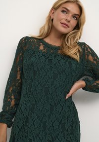 Robe en dentelle verte à manches longues, ornée d'un motif floral, avec un décolleté festonné et des détails transparents sur les manches et le corsage.