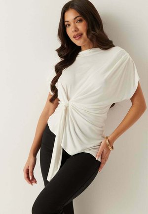 Kvinna med långt vågigt hår som bär en vit knuten blus och svarta byxor, poserar mot en enkel ljus bakgrund.