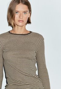Giovane donna con capelli corti castani, che indossa una maglietta a maniche lunghe beige e nera a righe, in piedi davanti a uno sfondo chiaro e uniforme.