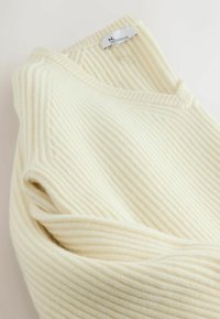 Cremefarvet ribstrikket sweater med V-hals, der har en blød tekstur og et mærke ved halsudskæringen.