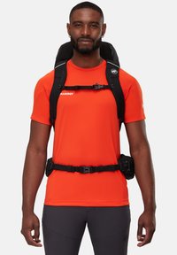 Svart ryggsäck med flera remmar över en starkt orange träningsskjorta. Texturerad tyg på skjortan, svart justerbar midjerem och synlig hårdvara.