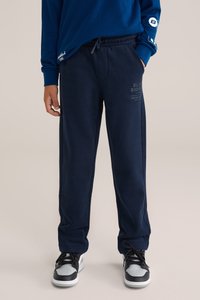 Jūras spēku sporta bikses ar brīvu piegriezumu, ar auklu jostas vietā, sānu kabatām un smalku teksta detalizāciju kreisajā kājā. Valkātas ar melniem un baltajiem sporta apaviem.