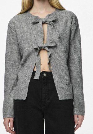 Personne portant un cardigan en maille gris avec deux nœuds à l'avant, partiellement ouvert pour révéler le ventre nu, associé à un pantalon noir taille haute.
