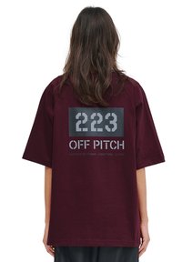 Burgunderröd bomulls t-shirt med korta ärmar. Baksidan har grå grafisk text: "223 OFF PITCH" och koordinater, på en svart rektangulär bakgrund.