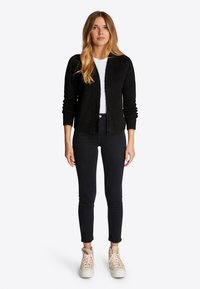 Cardigan nero lavorato a maglia con maniche lunghe, apertura frontale e polsini a coste. Indossato sopra una maglietta bianca e abbinato a jeans skinny neri e sneaker chiare.