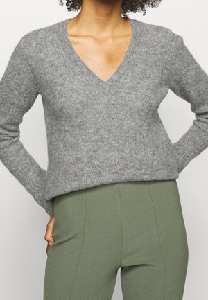 Femme portant un pull gris en V douillet et un pantalon taille haute vert olive, debout les mains sur les hanches sur un fond uni.