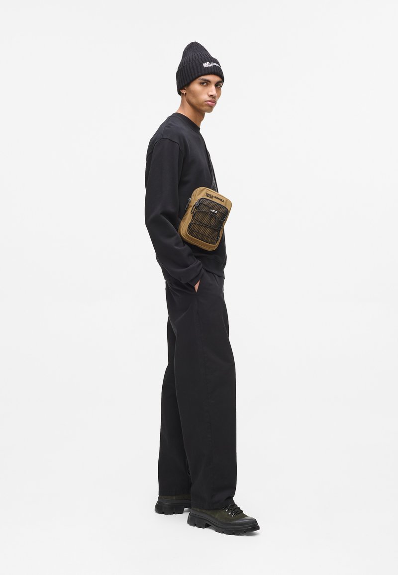 Schwarzer Pullover und Hose mit lockerer Passform, schwarze Mütze und eine braune Umhängetasche mit einem netzartigen Fach und Reißverschluss. Strukturierter Stoff.