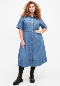 Denim-paitapuseromekko lyhyillä hihoilla, nappikiinnitys edessä, kaksi rintataskua ja gathered-alareuna. Yhdistetty mustien polvipitkien saappaiden kanssa.