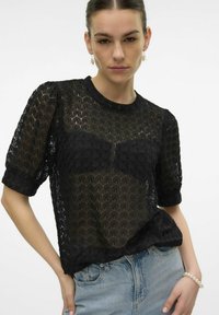 Top corto de encaje negro con mangas cortas que presenta un patrón geométrico, un escote redondo y una tela semitransparente. Combinado con jeans celestes.