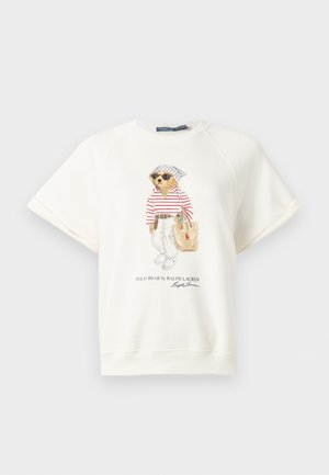 Γυναίκα με λευκό μπλουζάκι με σχέδιο και μπλε τζιν παντελόνι με φαρδιά πόδια, κρατώντας καφέ τσάντα, στέκεται μπροστά σε μονόχρωμο φόντο.