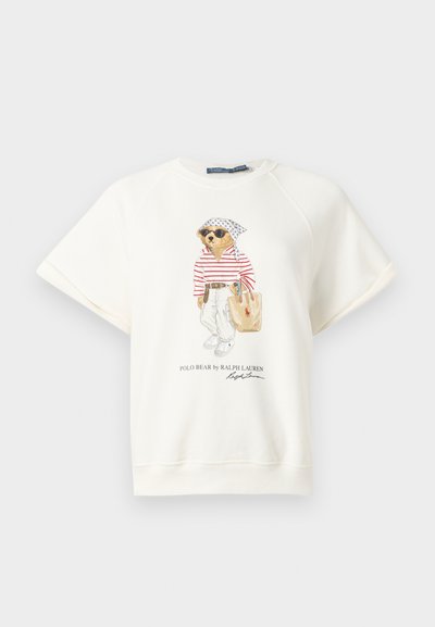 Λευκό φούτερ με κοντά μανίκια με μια αρκούδα που φοράει γυαλιά ηλίου, ριγέ πουκάμισο, λευκό παντελόνι, μαντήλι στο κεφάλι και κρατά τσάντα tote, τυπωμένο στο μπροστινό κέντρο.