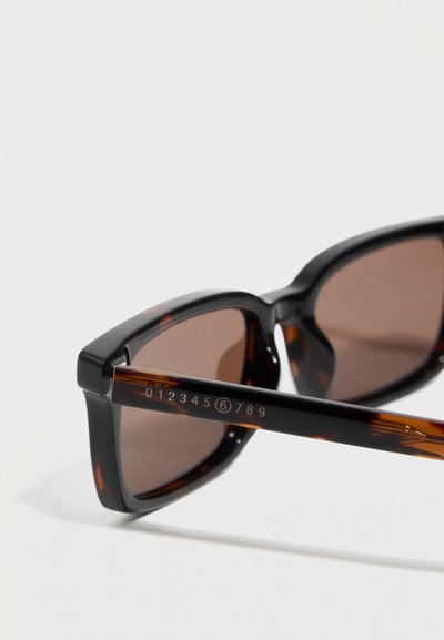 Γυαλιά ηλίου με ταρταρούγα και σκουρόχρωμους καφέ φακούς, που δείχνουν τον αριστερό βραχίονα με αριθμούς 012345 (6) 789 σε λευκό χρώμα.