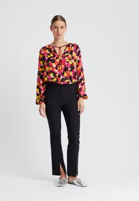 Bunte Langarmbluse mit Blumenmustern in Rosa, Gelb und Schwarz, kombiniert mit schwarzen, taillierten Hosen mit seitlichen Schlitzen und silbernen Slippers.