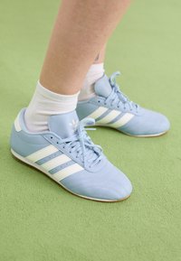 Jasnoniebieskie buty sportowe wykonane z materiału syntetycznego, z białymi paskami i sznurówkami, o teksturowanym wzorze i gumowej podeszwie.