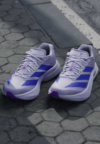 Ζευγάρι λευκά και μωβ παπούτσια για τρέξιμο Adidas Adizero, τοποθετημένα σε γκρι πλακόστρωτο με εξαγωνικά πλακάκια κοντά σε πεζοδρόμιο δρόμου.