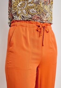 Pantalons orange à cordon de serrage en tissu lisse, dotés de poches latérales et d'une coupe décontractée. La taille est élastique pour plus de confort.