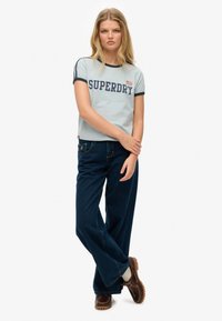 Hellblaues kurz geschnittenes T-Shirt mit marineblauen Akzenten und dem Schriftzug „SUPERDRY“ in fetten Buchstaben. Kombiniert mit weitem dunkelblauen Jeans und braunen Schuhen.