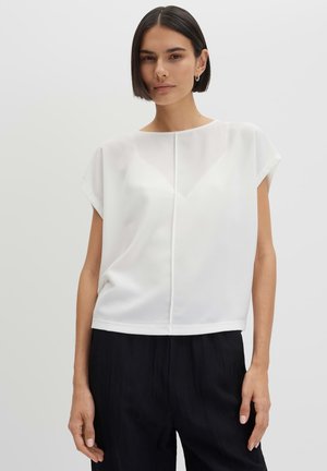 Vrouw met kort donker haar, een wit blouse met korte mouwen en een zwarte broek, staand tegen een effen lichte achtergrond.