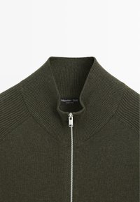 Knit, olive green sweater featuring a high collar and full zipper. Textured knit pattern on shoulders and snug fit. Brand label visible inside.