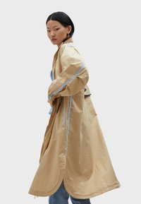 Beige long coat made of lightweight fabric with blue accents, featuring a relaxed fit, side pockets, and a curved hem.