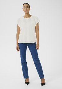 Haut blanc à manches courtes avec un motif en œillets, associé à un jean bleu. Le modèle porte des escarpins noirs à bout pointu et se tient sur un fond neutre.
