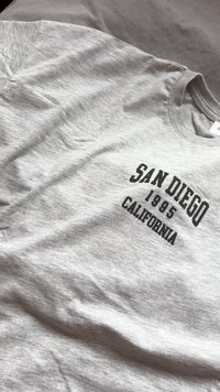 Tricou din bumbac gri, cu text negru care spune „SAN DIEGO 1995 CALIFORNIA.” Design clasic cu guler tip crew și o textură fină, ușoară.