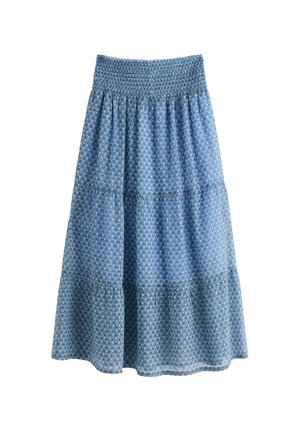 Nuori nainen seisoo valkoisessa neulotussa neuletakissa, vaaleansinisessä topissa, pitkässä sinisessä kuvioidussa hameessa ja vaaleanpunaisissa kukkakuvioisissa tohveleissa yksiväristä taustaa vasten.