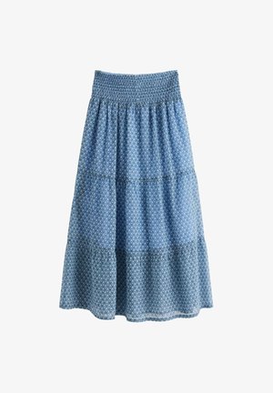 Nuori nainen seisoo valkoisessa neulotussa neuletakissa, vaaleansinisessä topissa, pitkässä sinisessä kuvioidussa hameessa ja vaaleanpunaisissa kukkakuvioisissa tohveleissa yksiväristä taustaa vasten.