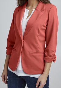 Blazer corail à manches trois-quarts, coupe cintrée, col cranté et deux poches avant. Superposé à une chemise blanche.