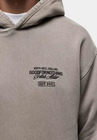 Grijze hoodie van zacht materiaal, met een geborduurd logo in donkergrijs. Het ontwerp heeft een capuchon met trekkoord en geribbelde mouwen.