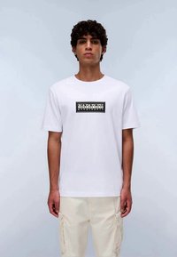 Jovem com cabelo encaracolado, vestindo uma t-shirt branca da Napapijri e calças cargo de tom bege claro, posando contra um fundo claro e neutro.