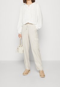 Blusa bianca a maniche lunghe con ricami sottili, abbinata a pantaloni beige chiaro a vestibilità ampia e a una piccola borsa bianca con cordoncino. Scarpe platform color beige.