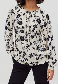 Schwarz blusiform mit floralen Mustern aus leichtem Stoff, mit rundem Halsausschnitt, gefältelten Details und langen Ärmeln. Kombiniert mit schwarzen Hosen.