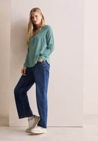 Camicia a maniche lunghe verde chiaro, con texture e colletto con bottoni, abbinata a jeans a gamba dritta blu scuro e sneakers bianche.